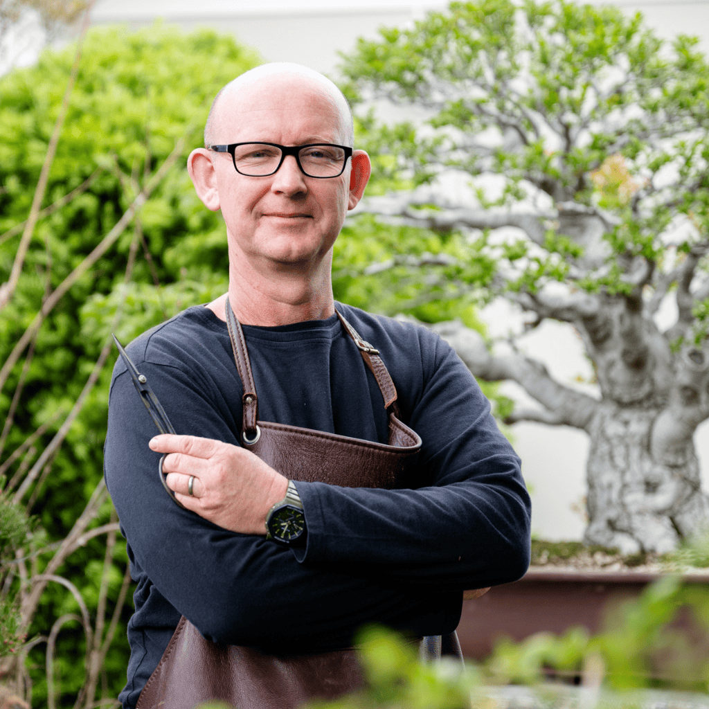 Man wearing glasses and an apron standing in a garden with trees in the background